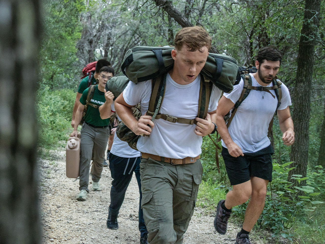 Marine candidates carrying heavy rucksacks during endurance march on wooded training trail.