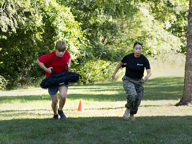 A recruit in a red shirt and their Marine Recruiter perform cardio exercises during Marine Corps physical training outdoors.