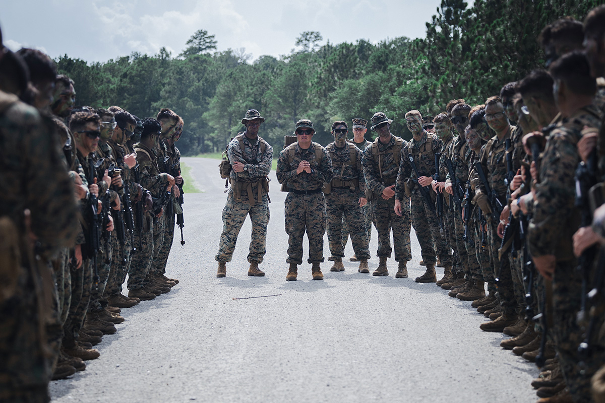 Officer briefs Marines gathered in field during outdoor training instruction under clear sky with pine trees.