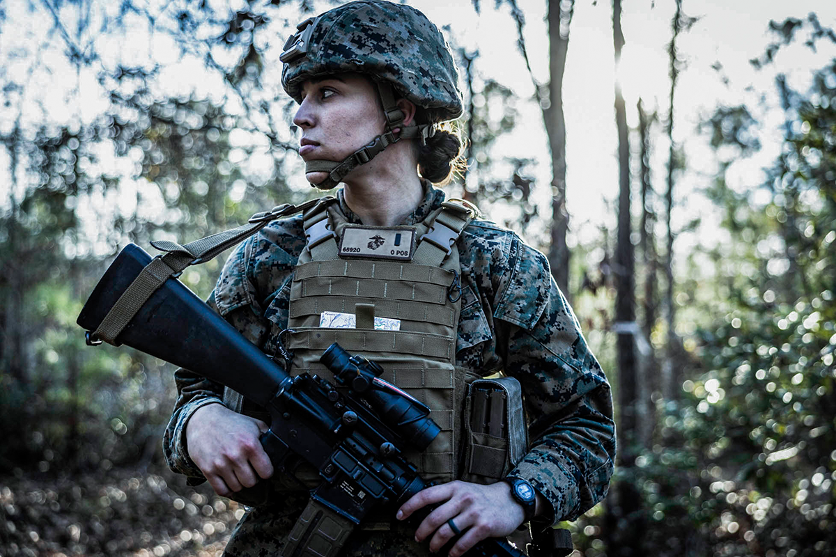 Officer in tactical gear with rifle stands alert during cyber field training exercise in wooded area at Camp Lejeune.