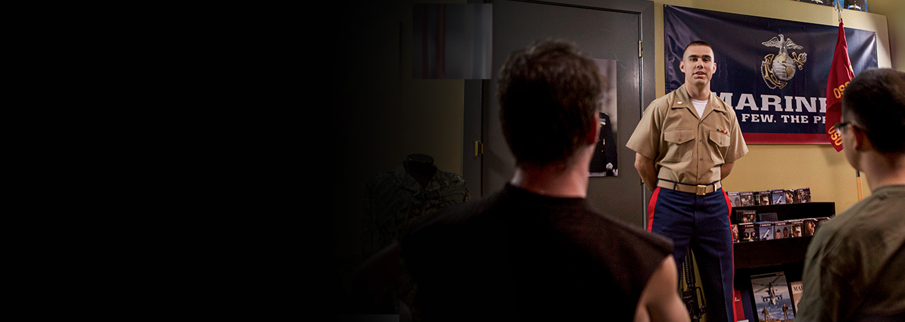 Recruiter in dress uniform speaks with prospective recruits at recruiting station beneath Eagle Globe and Anchor emblem.