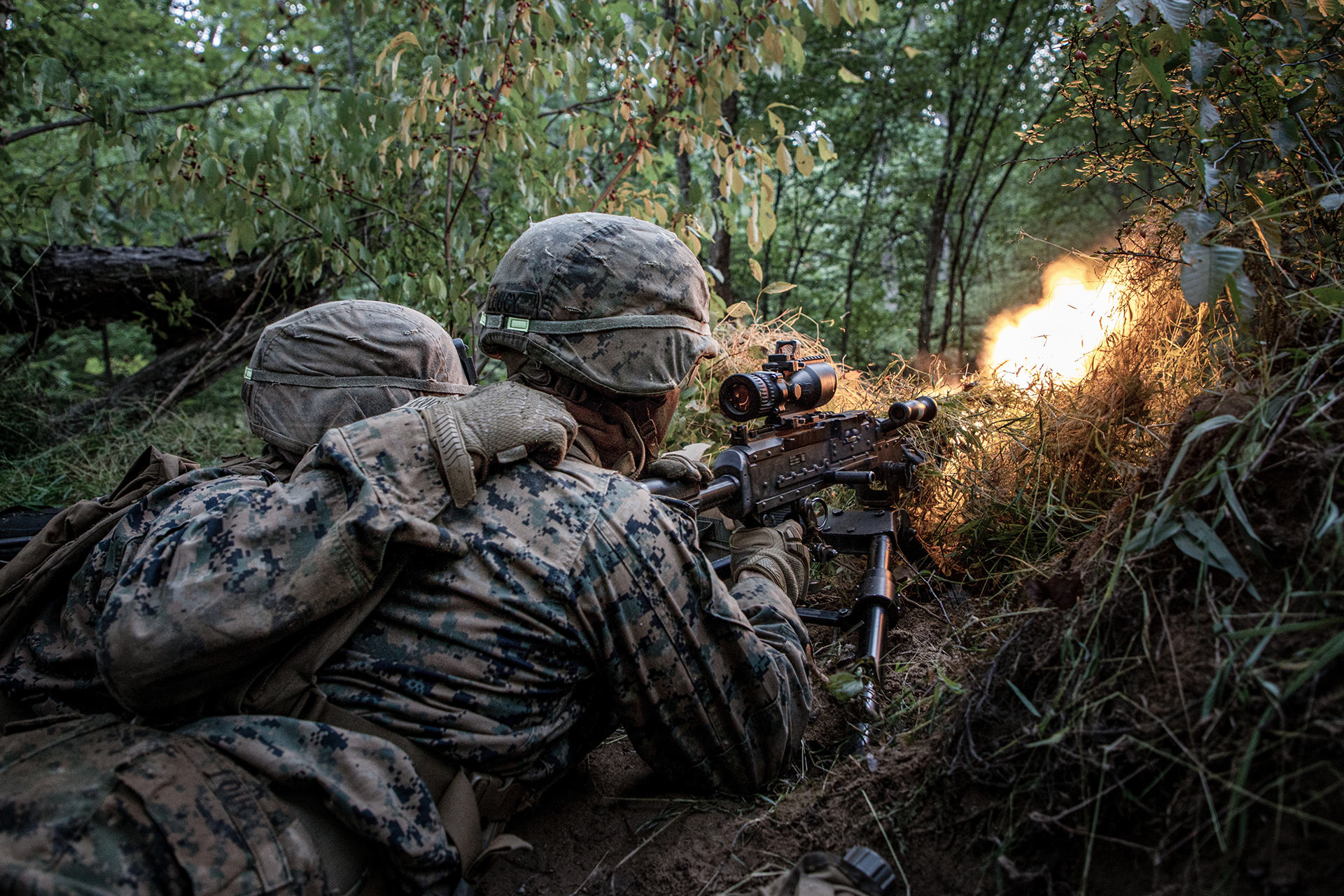 Marine officers firing machine gun with muzzle flash during Phase 3 rifle platoon commander training in wooded environment.
