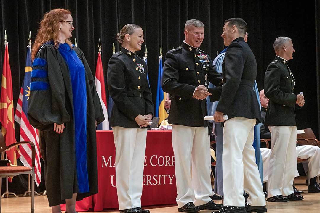 Marines in dress blues shaking hands with another Marine. Marine Corps University banner in background at a college graduation ceremony.