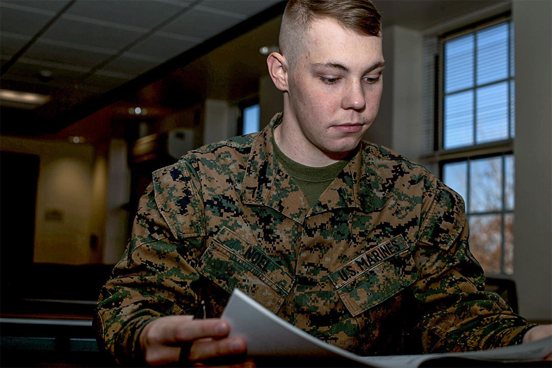 Marine in uniform at a desk in a classroom, reading a document.