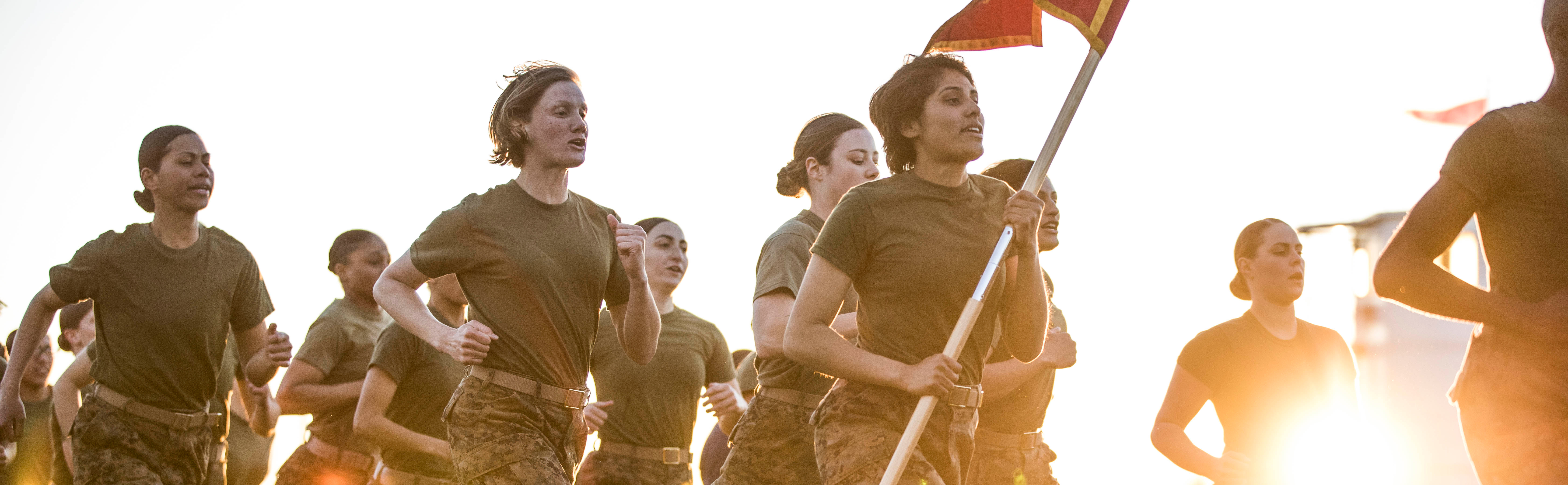 A group of female Marines run together in the early morning. One of the Marines carries a guidon. 