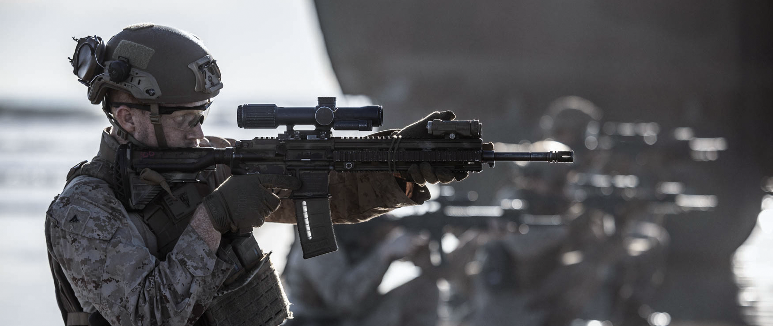 A Marine, in profile, looks down the scope of his rifle. He is on a beach with other Marines. They just got off an amphibious vehicle. 