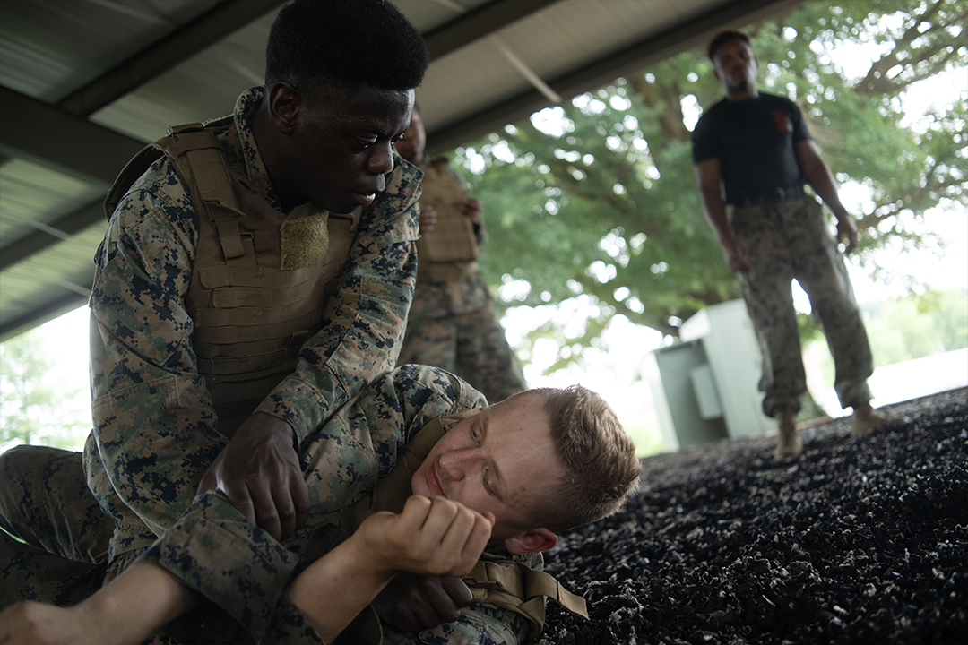 Two Marine recruits wrestle on the ground. One pins the other. A drill instructor oversees the training.