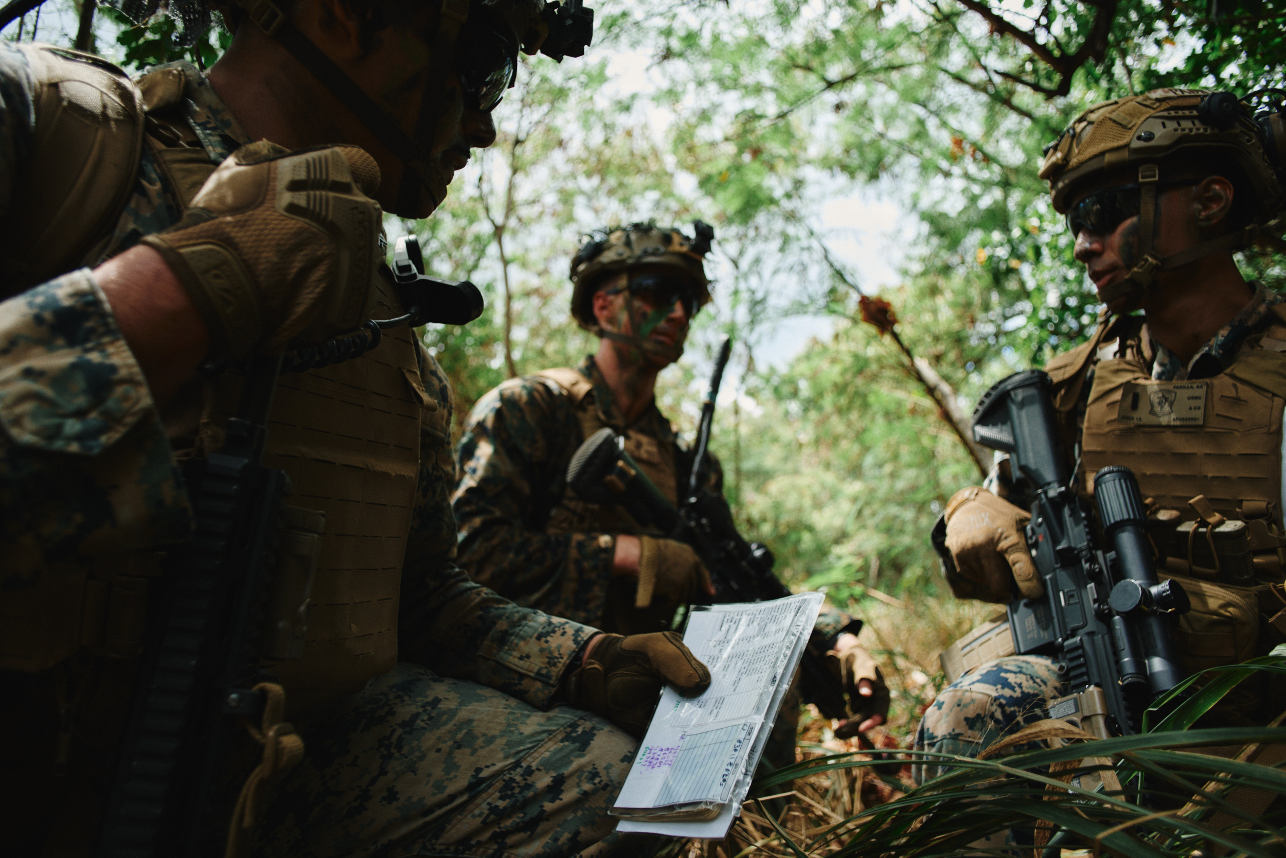 Marines in camouflage face paint kneel together in dense woods, reviewing a map as they plan their next move.