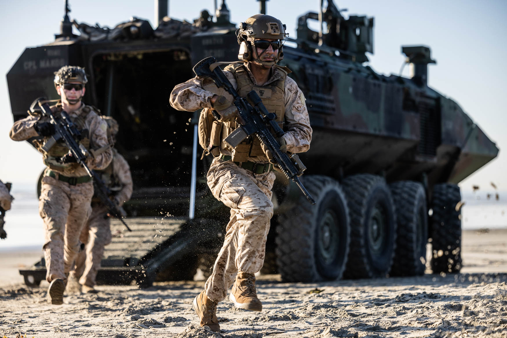 Marines advance on foot across a beach in front of an amphibious combat vehicle.