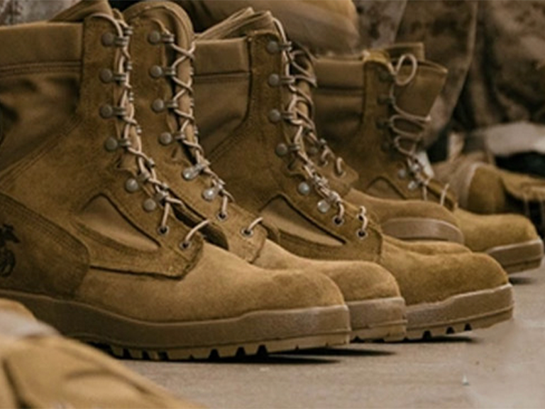 Multiple pairs of tan Marine combat boots with laces, one featuring the Marine Corps emblem, lined up on a dark surface.