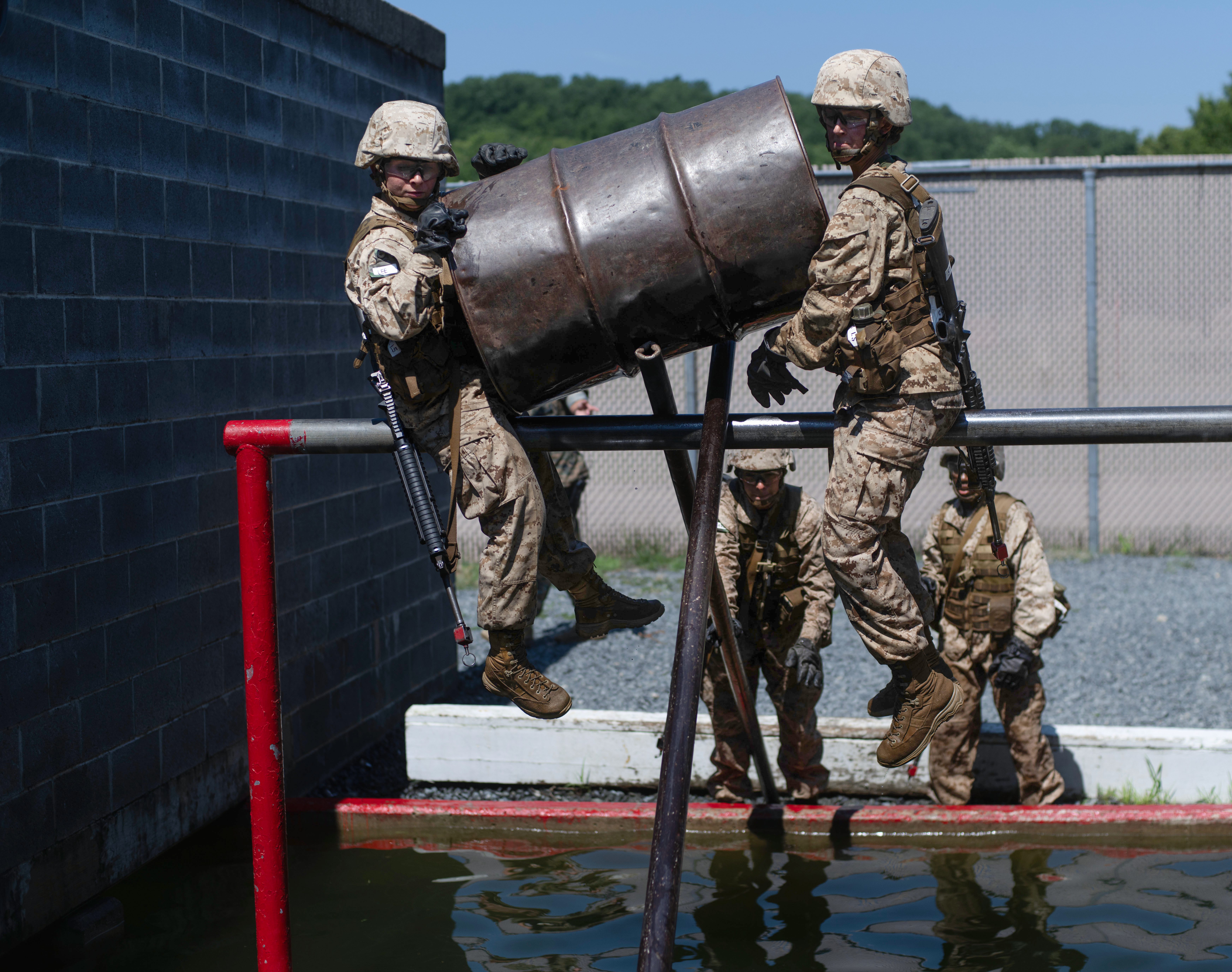 Two Marine officer candidates carry a heavy barrel across a metal rail over a water pit as teammates wait to take it, working through a Leadership Reaction Course obstacle.