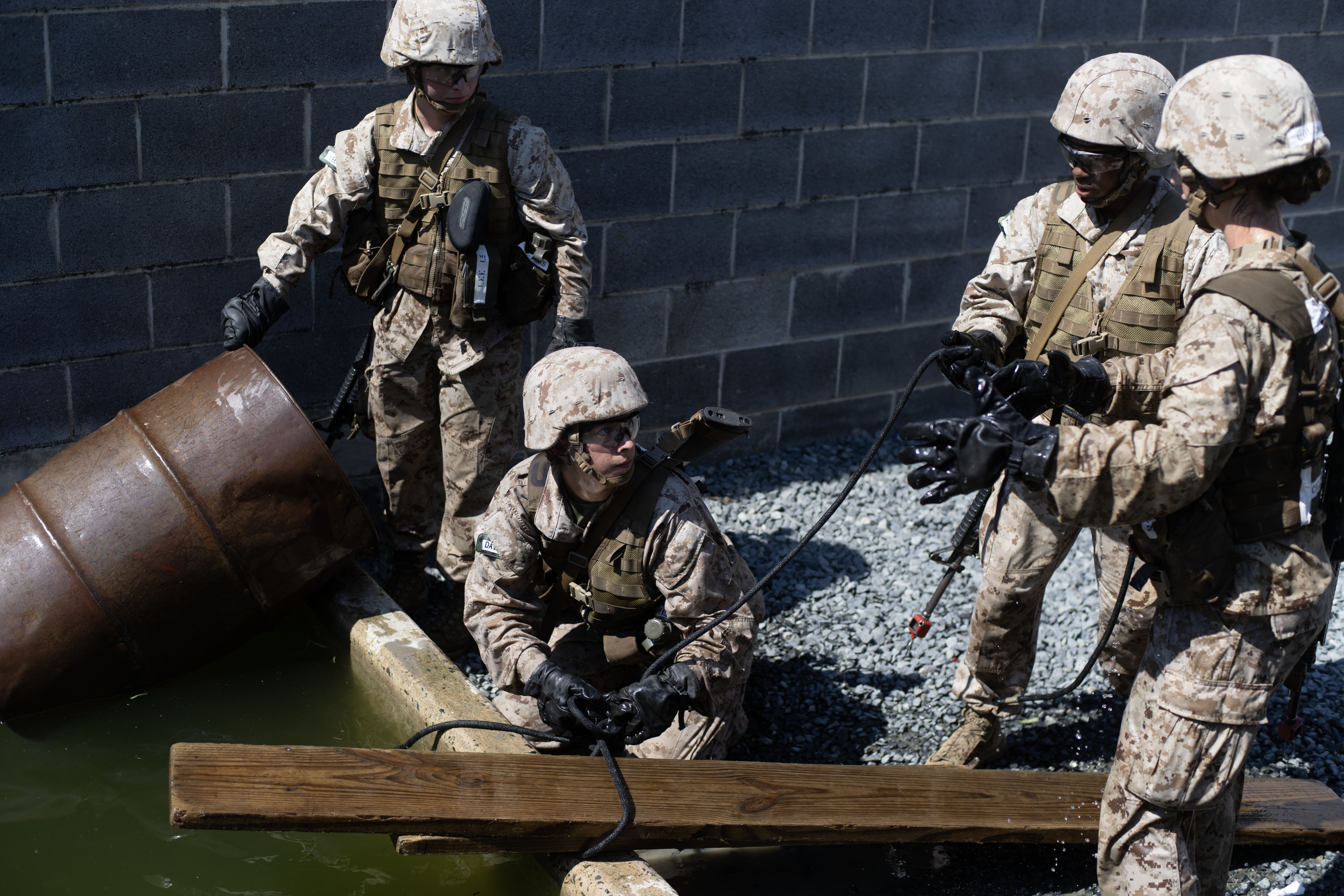 Marine officer candidates in camouflage utilities and helmets work together to move a rope across a barrel-and-plank water obstacle at the Leadership Reaction Course.