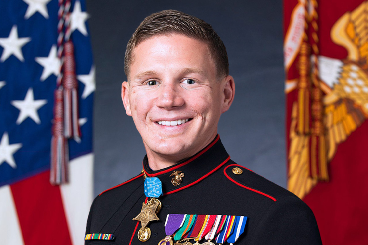 Kyle Carpenter, Medal of Honor recipient Marine, in dress blue uniform with American and Marine Corps flags.