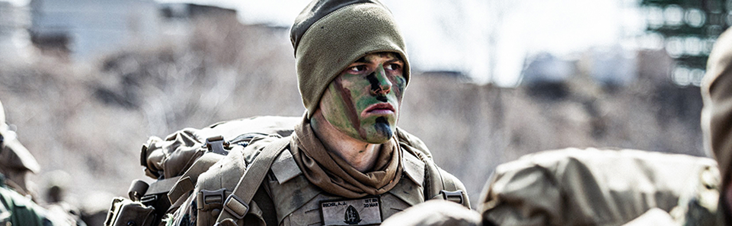 Marines in camouflage face paint and gear march in formation carrying packs during training exercise.