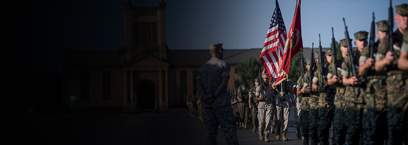 Marines walk in file at a USMC graduation ceremony. They are dressed in camouflage, carry rifles, and one holds USA and USMC flags.
