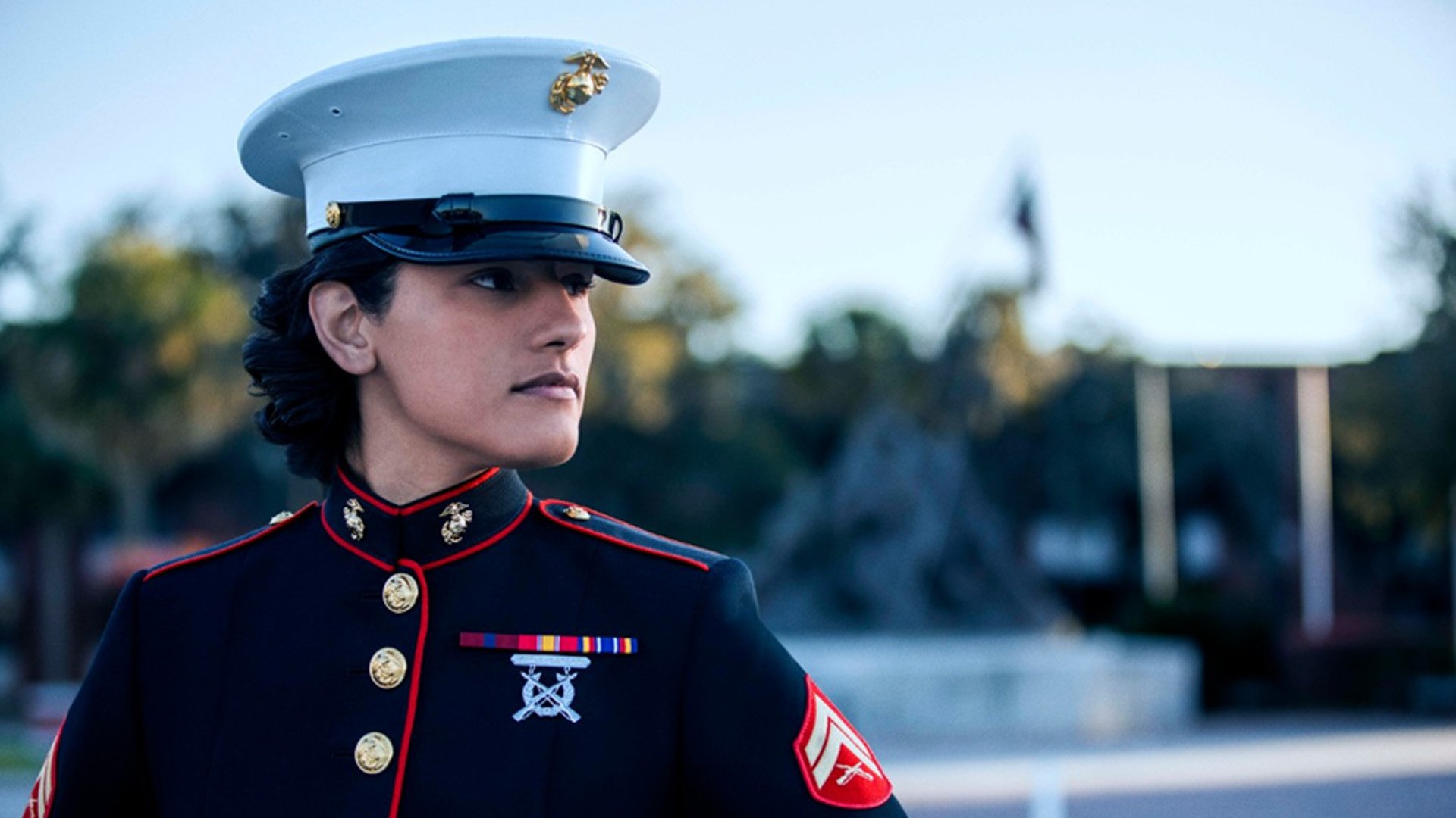 Marine in blue dress uniform with red trim, gold buttons, ribbons, and white hat looks right, against blurred outdoor background.