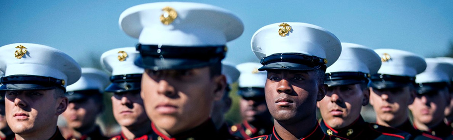 Close-up of a line of Marines in blue dress uniforms and white hats, looking forward under a clear sky.