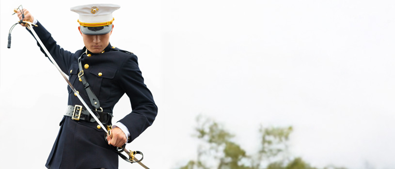 A Marine in dress uniform sheathing a mameluke sword, looking down, with a blurry outdoor background.