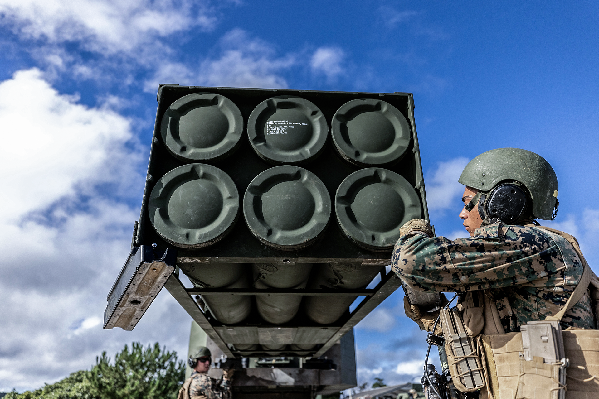 Marine inspects HIMARS rocket artillery system launcher pod with six missile tubes under clear blue sky.
