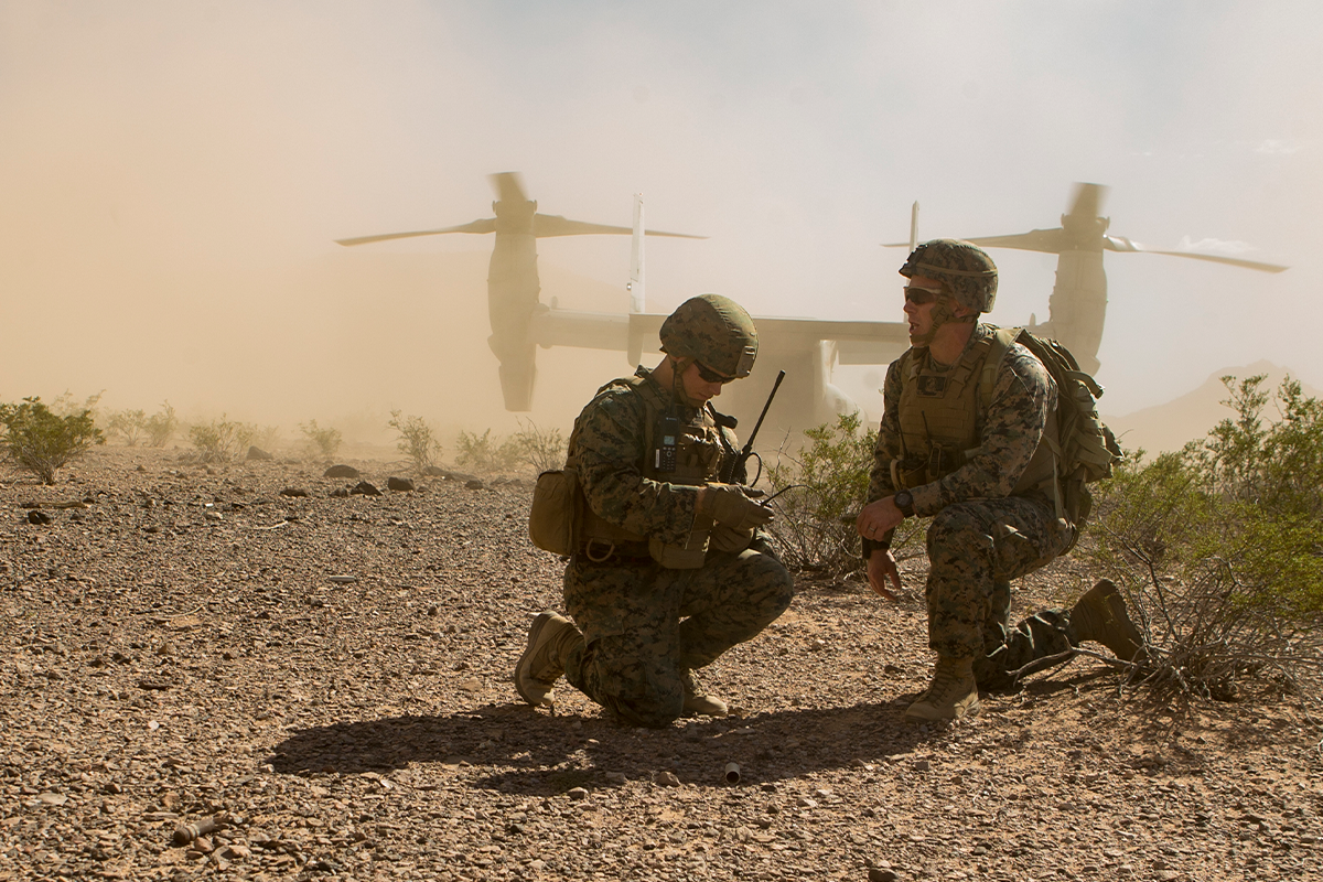 Marines communicate via radio as V-22 Osprey aircraft lands during Marine Expeditionary Force training in dusty conditions.