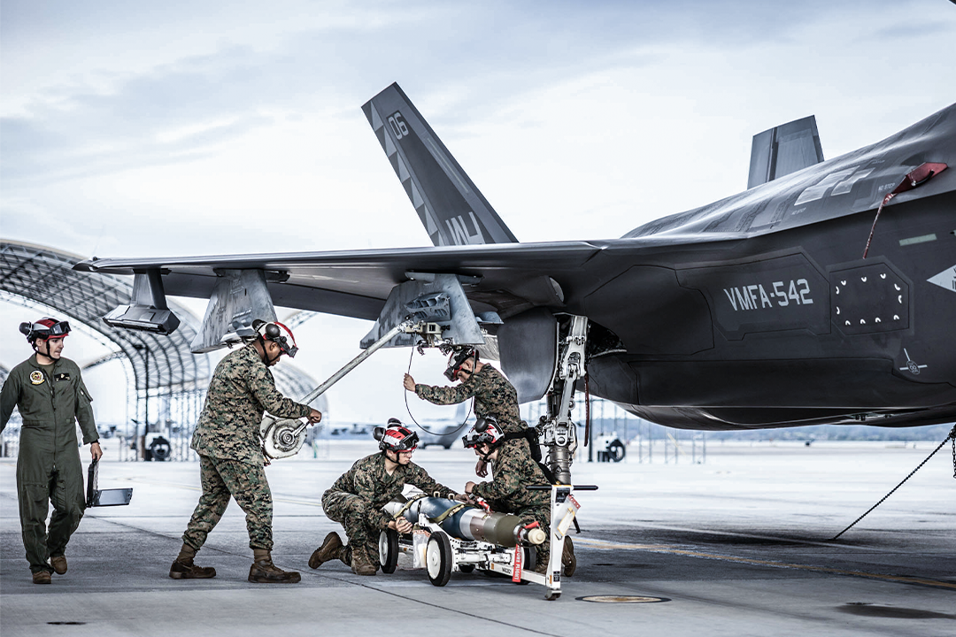 Marines in camo and flight suits load a missile onto the wing of a F-35B on an airfield tarmac. 