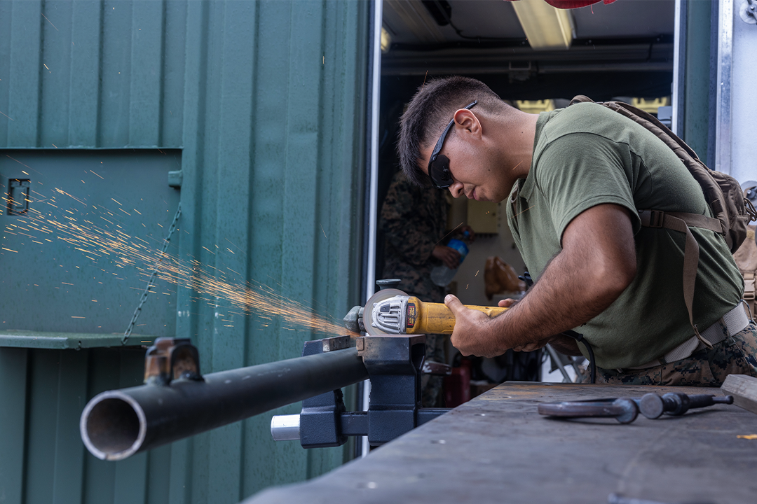 A machinist Marine works on metal pipe with an orbital sander. Sparks fly away from the work piece. 