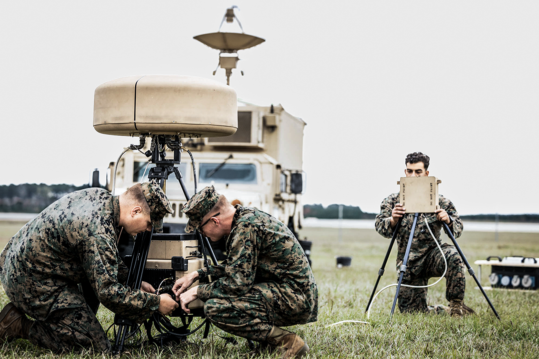 Three intel and planning Marines set up equipment in a grassy field.