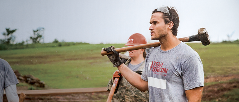 Jake Wood carrying sledgehammer during Team Rubicon disaster relief volunteer work in field.