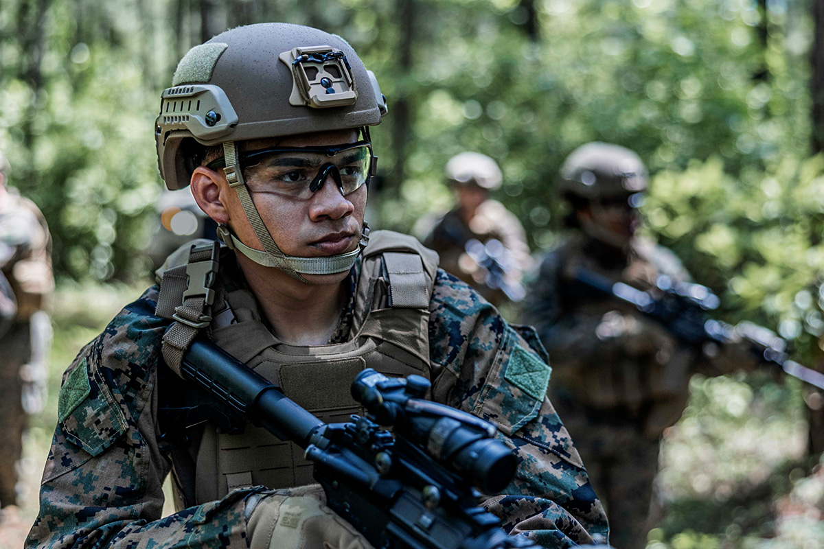 An infantry Marine walks through a forest along with the rest of his squad. They have on full gear and have their rifles at the ready.