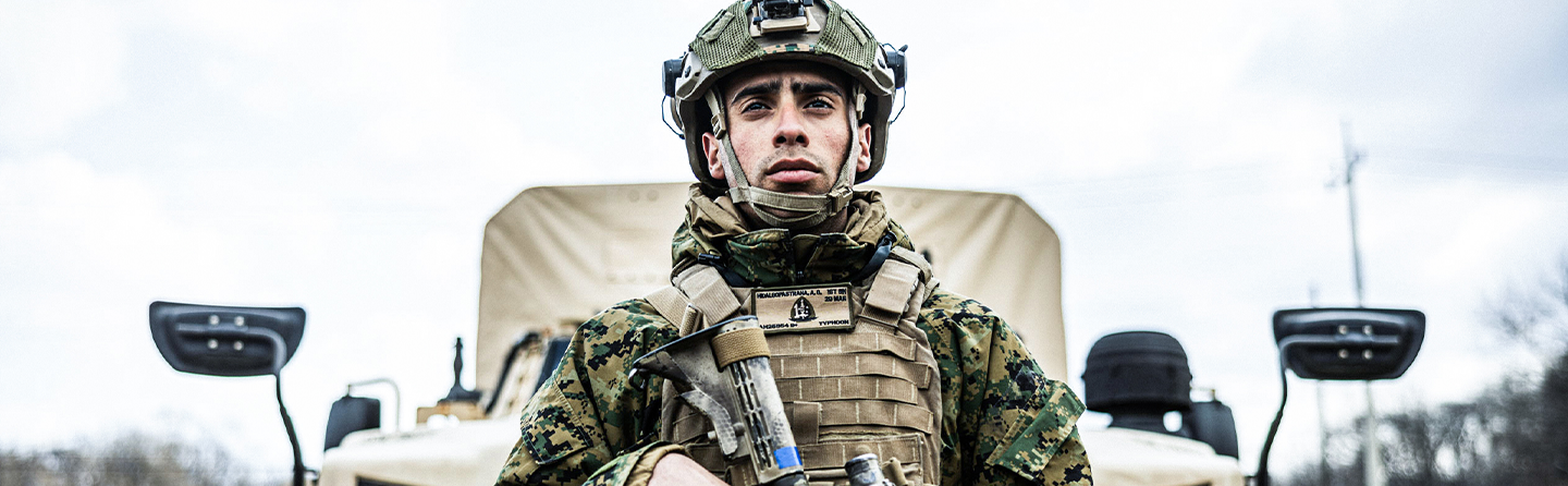 An infantry Marine stands in front of a transportation truck. He has a combat helmet, rifle, body armor, and carries a rifle with scope.