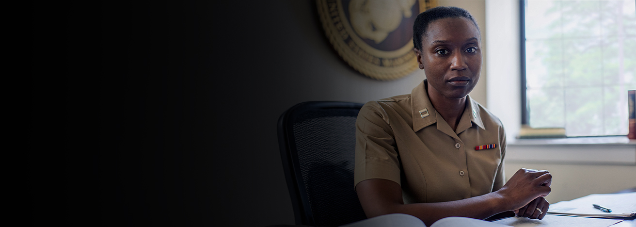 A Judge Advocate sits at her desk in her office. Work is spread out before her and a large Eagle Globe and Anchor plaque is behind her.