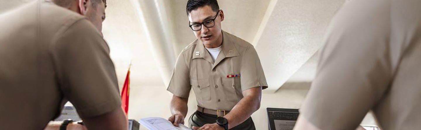 Three Marine Judge Advocates gather around a table in a courtroom. One is showing a document to the other Marines. 