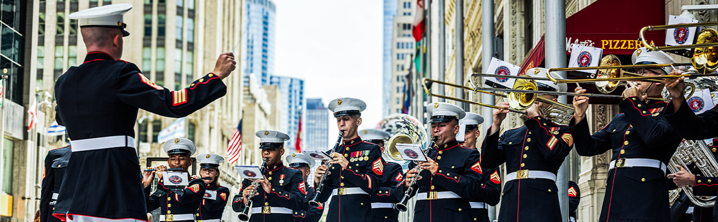 Marine band performs in dress uniforms on a city street, conductor leading musicians with brass and percussion instruments.